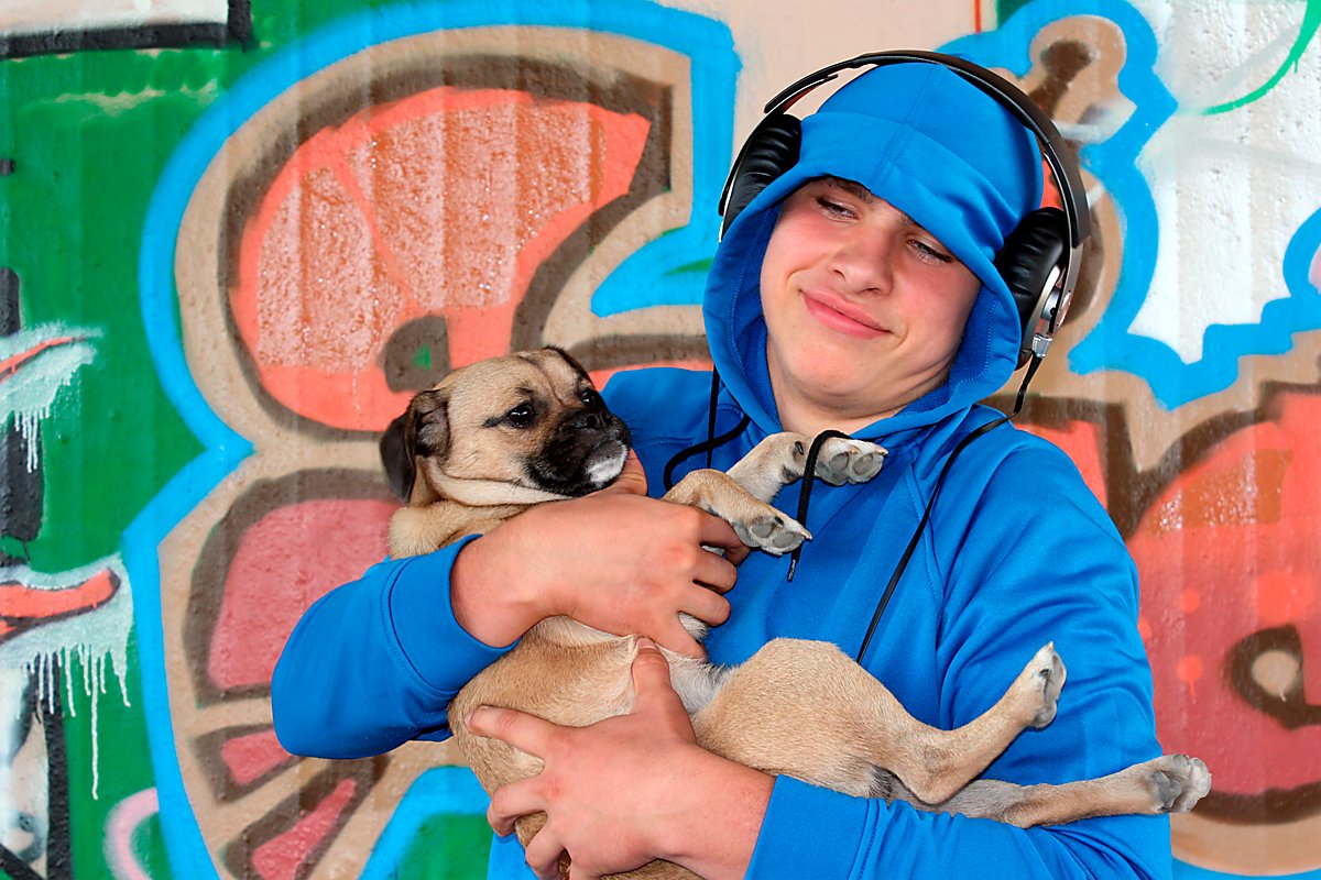 Man sieht einen Jugendlichen mit Kopfhörern auf den Ohren und einem Welpen in den Armen vor einer Graffiti-Wand.