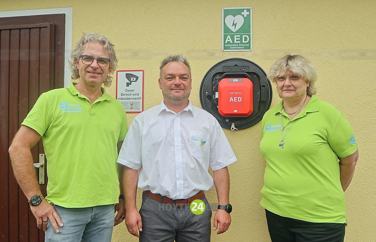 Man sieht Heiko Keller vom Heimat- und Kulturverein Lohsa sowie Katrin Schütz und Thomas Zentsch vom Sanitätshaus Zentsch in Bautzen vor einem neuen Defibrillator in Lohsa.