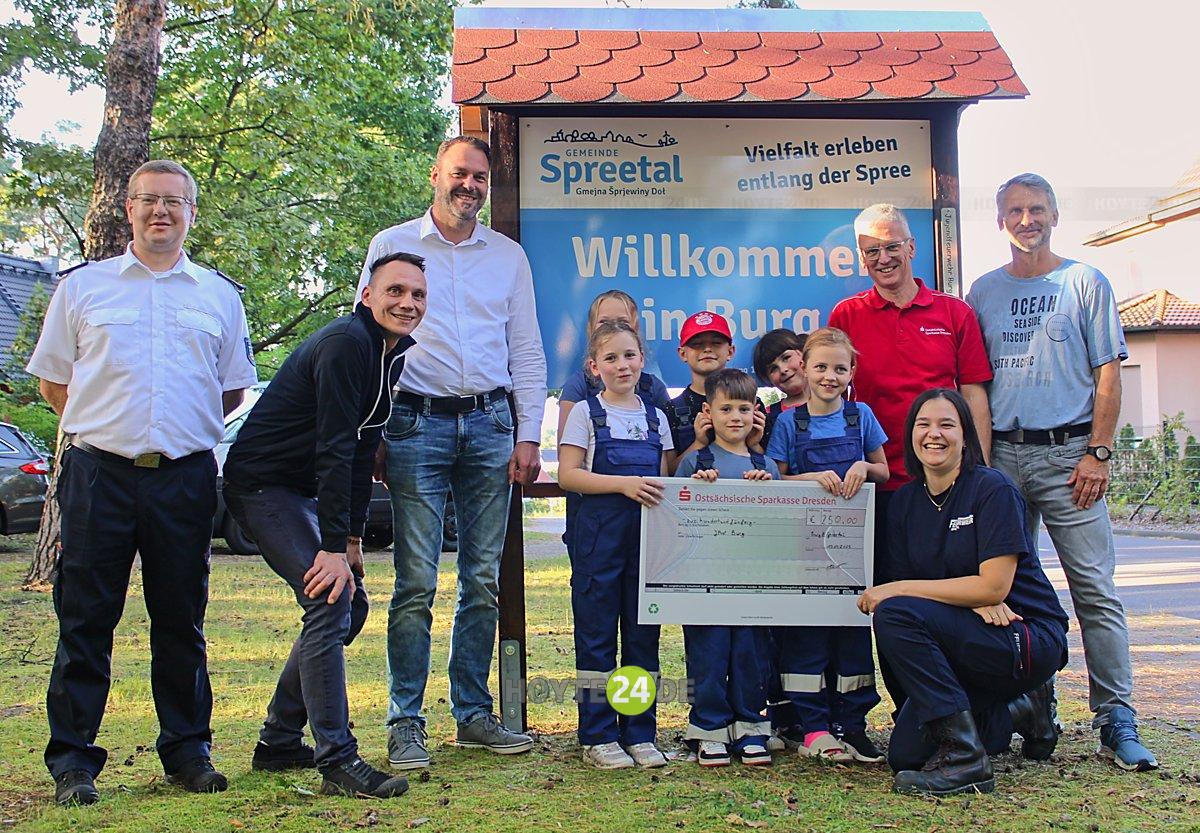 Man sieht die Kinder und ein paar Erwachsene mit dem Preisgeld-Scheck vor einer der beiden neuen Willkommenstafeln.