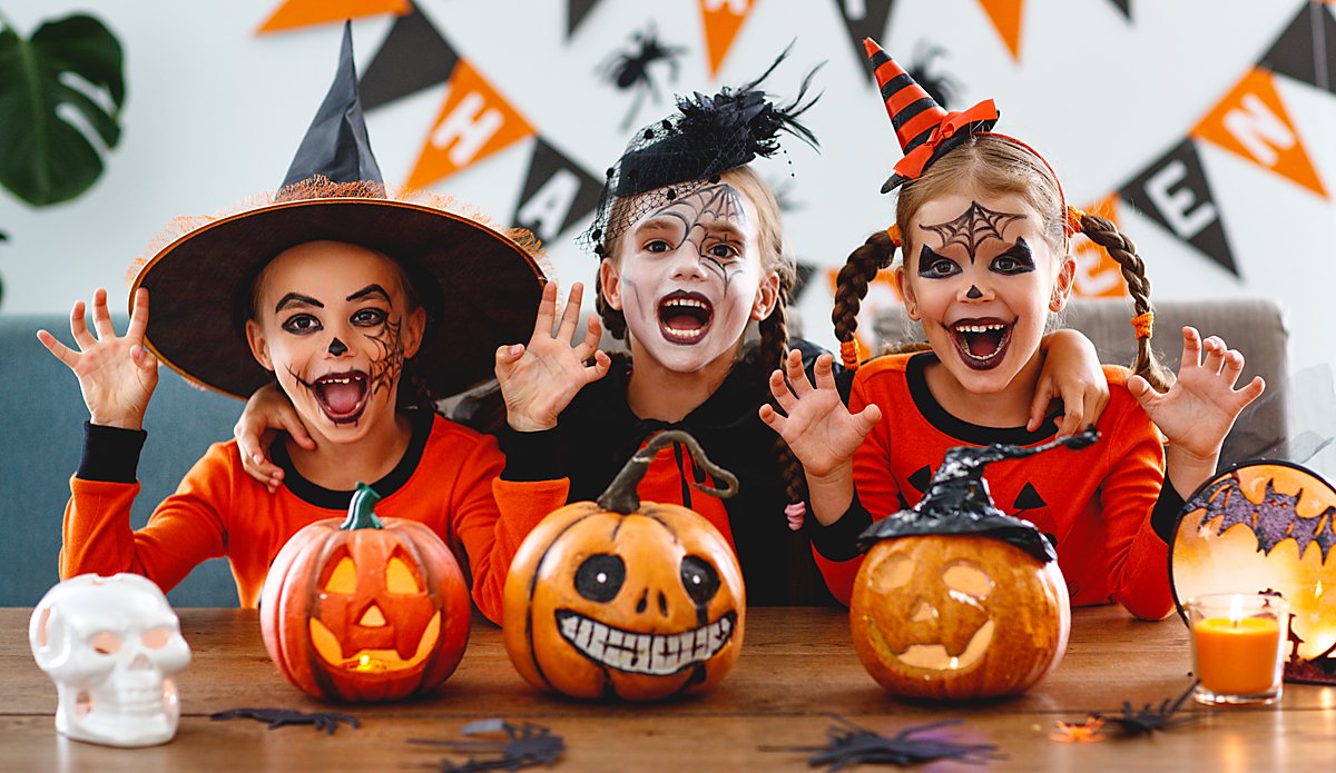 Man sieht Kinder mit Halloween-Kostümen und drei Kürbisse, in die Gesichter geschnitten sind.