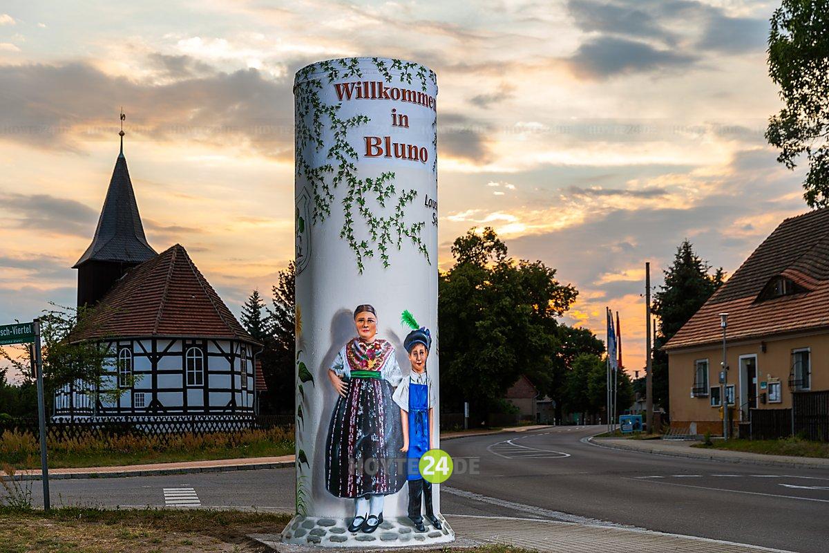 Man sieht die Litfaßsäule mit Willkommensgruß und Menschen in sorbischer Tracht in Bluno.