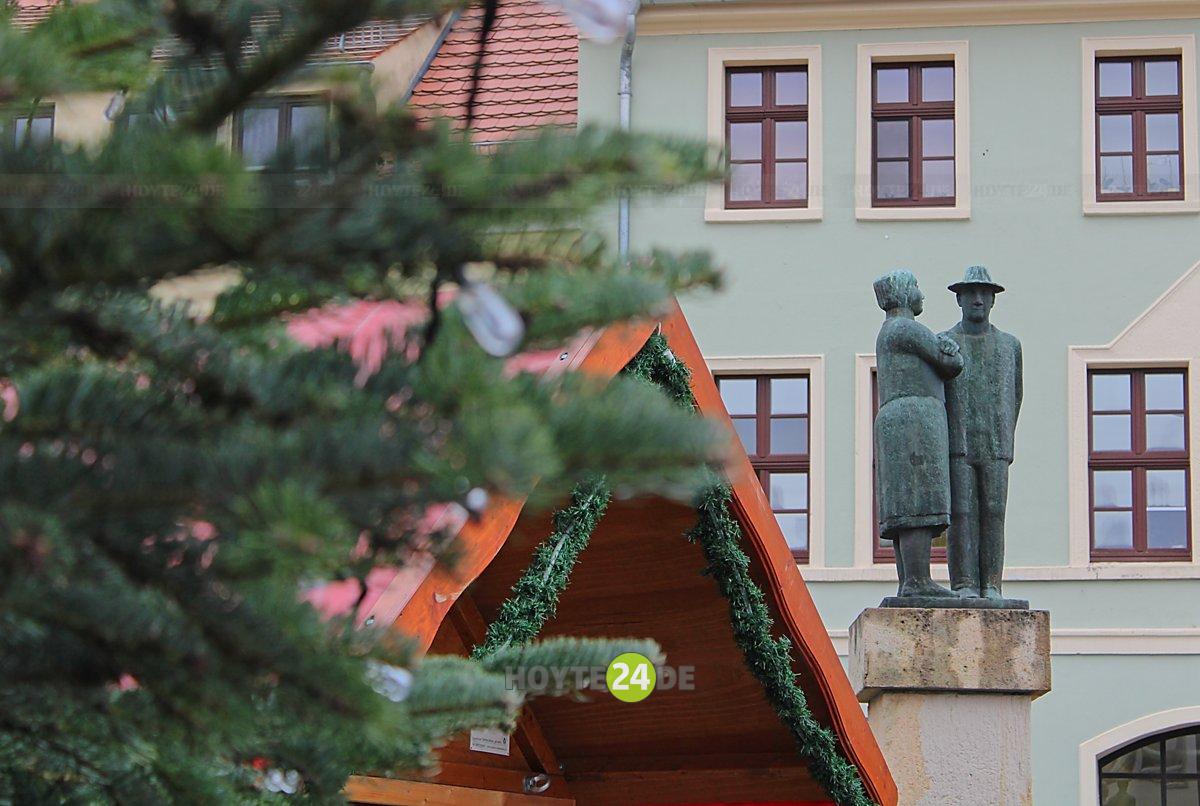 Man sieht Grün vom Weihnachtsbaum auf Hoyerswerdas Markt, das Dach einer Weihnachtsmarkt-Hütte sowie das Paar vom Sorbenbrunnen.