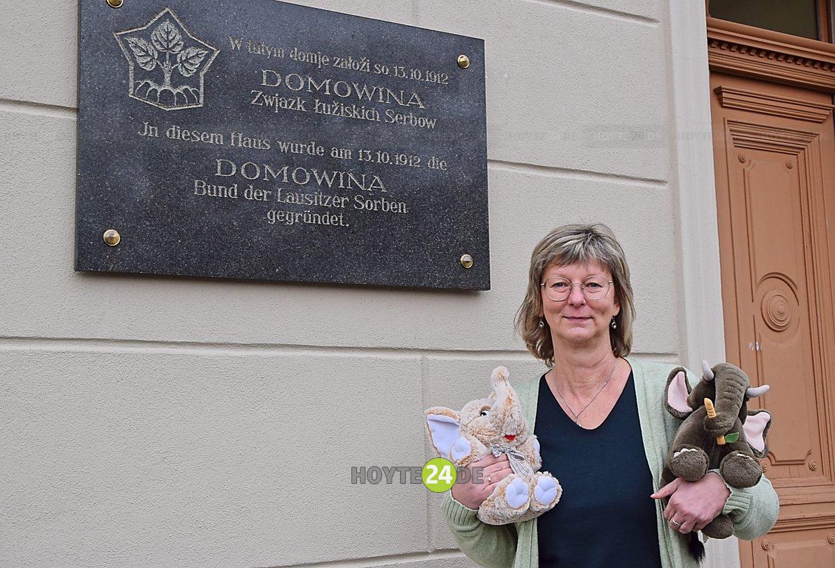 Man sieht Ina Züchner mit zwei Plüsch-Elefanten vor der Tafel "In diesem Haus wurde am 13.10.1912 die Domowina Bund Lausitzer Sorben gegründet.